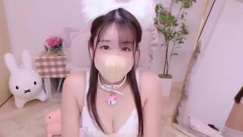 Media: Video of an East Asian woman in a white bunny costume with a fluffy tail, wearing a face mask, kneeling on a soft carpet in a pastel-themed room with bunny decor.