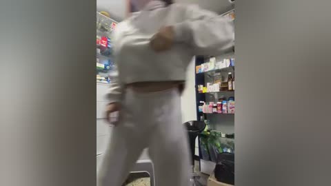 Video of a person in white athletic clothing, blurred, standing in a cluttered pharmacy with shelves of products and plants.