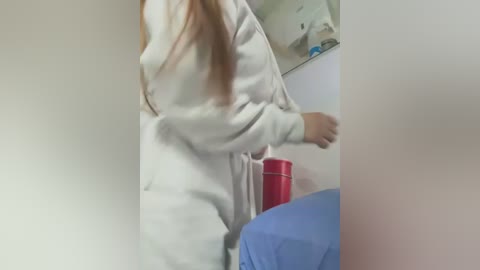 Video of a woman in a white bathrobe, leaning forward, with long, straight, light brown hair. Background shows a red plastic cup on a blue cloth surface and a blurred wall.