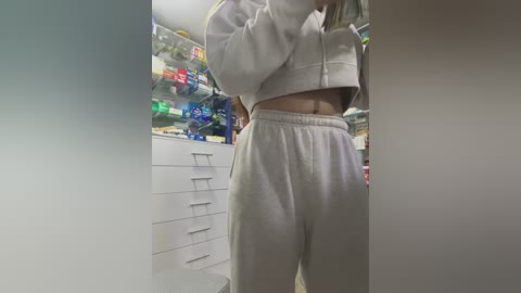 Video of a woman in a beige hoodie and matching sweatpants, standing in a cluttered room with shelves of colorful snacks and a white dresser in the background.