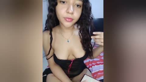 Media: Video of a young Asian woman with curly hair, wearing a black lace bra, sitting on a bed with a red and white striped blanket, taking a selfie.