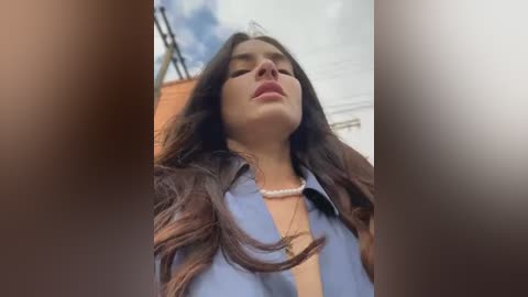 Media: Video of a young woman with long dark hair, wearing a blue shirt, standing outdoors under a cloudy sky, looking up with a serene expression.