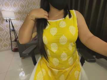 Media: Video of a woman with long black hair, wearing a sleeveless yellow dress with white floral patterns, seated indoors, with a beige wallpaper and a black lamp in the background.