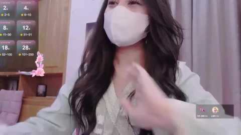 Media: Video of an Asian woman with long black hair, wearing a white mask, light green sweater, and a pink bracelet, in a dimly lit room with a TV displaying a weather forecast.