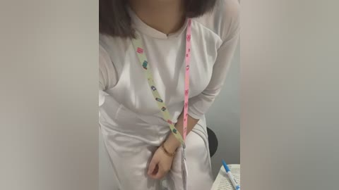 Media: Video of a woman with medium skin tone wearing a white, long-sleeved shirt, holding a lanyard with a colorful pattern. She stands indoors, partially blurred, near a desk with a notebook and pen.