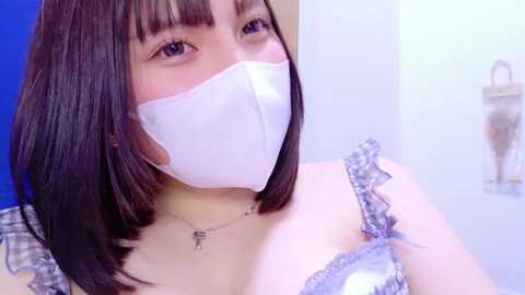 Media: Video of a young Asian woman with straight, shoulder-length dark brown hair, wearing a white surgical mask and a light blue, frilled, off-shoulder top. Background is a bright blue wall and a white shelf with a small, brown, cylindrical object.