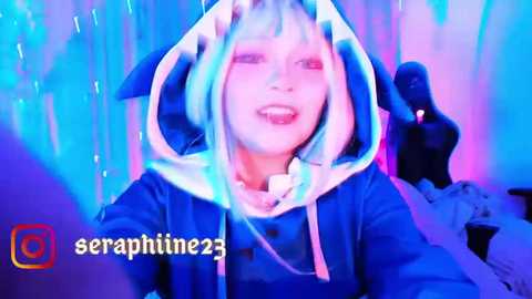 Media: A vibrant video of a young woman with pale skin and blue hair, wearing a blue and white hooded cosplay outfit, surrounded by neon blue and purple lighting.