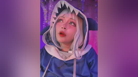 Media: Video of a young woman with pale skin and white hair, wearing a blue shark hoodie with a shark mouth hood, looking up in a purple-lit room.