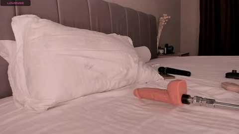 Media: Video of a neatly made bed with a white pillow and beige headboard. Various sex toys, including a pink dildo, black vibrator, and a screwdriver, are scattered on the bedspread.