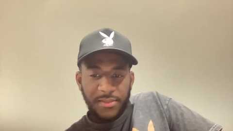 Media: Video of a young Black man with a short beard, wearing a black Playboy cap and grey t-shirt, against a plain beige background.