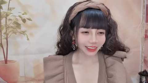 Media: Video of an East Asian woman with long black hair, wearing a beige headband and dress, smiling against a blurred, beige wall background with a potted plant.