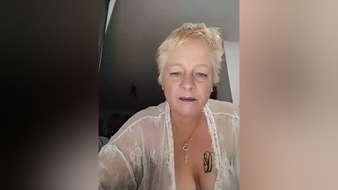 Media: Video of a middle-aged woman with short blonde hair, wearing a sheer white lace robe that reveals her cleavage, and a gold necklace with a pendant. She's indoors, with blurred background.