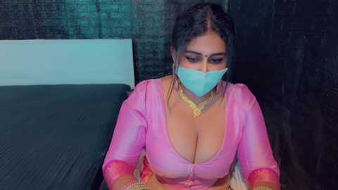 Media: Video of a woman in a pink, low-cut top, with a blue mask, sitting on a bed with a dark headboard, wearing a gold necklace.