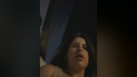 Media: Video of a woman with fair skin, dark hair, and closed eyes, lying on a dark surface, possibly a bed, with a dimly lit room in the background.