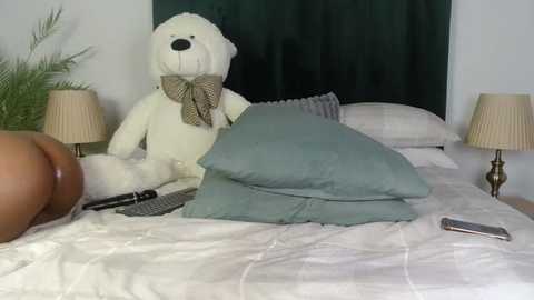 A video of a plush white teddy bear with a bow tie, resting on a neatly made bed with a grey pillow and beige lampshades on each side.