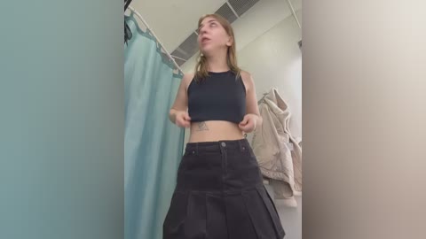 Media: Video of a young Caucasian woman with light skin and shoulder-length blonde hair, wearing a black crop top and high-waisted black skirt, standing in a brightly lit, simple changing room with a teal curtain and white walls.