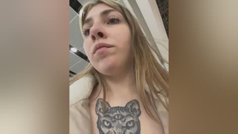 Media: Video of a young woman with fair skin and long blonde hair, wearing a beige robe. She has a large, intricate tattoo of a feline's face with three eyes on her chest. Background shows a modern room with white walls and a ceiling light.