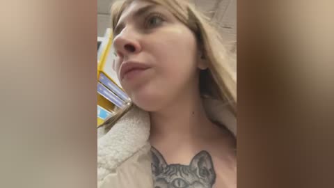 Media: Video of a blonde woman with fair skin, wearing a beige jacket, showing a detailed grey cat tattoo on her chest. Background shows blurred office items.