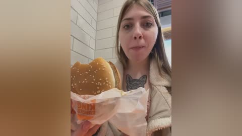 Media: Video of a young woman with light skin, long brown hair, and a tattoo on her chest, holding a McDonald's sandwich, seated in a white-tiled fast-food restaurant.