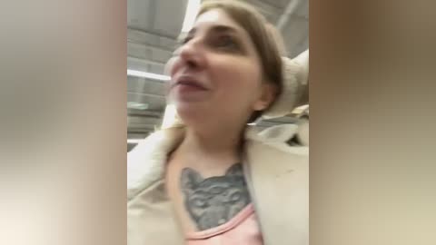 Media: A blurry video of a woman with light skin and brown hair, wearing a beige coat over a pink shirt with a gray, detailed tattoo on her chest. She has a neutral expression. The background shows a tiled wall and metallic surfaces.