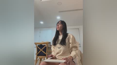 Media: Video of a young woman with long black hair, wearing a cream sweater, sitting on a wooden chair in a modern, minimalist room with white walls and recessed lighting.