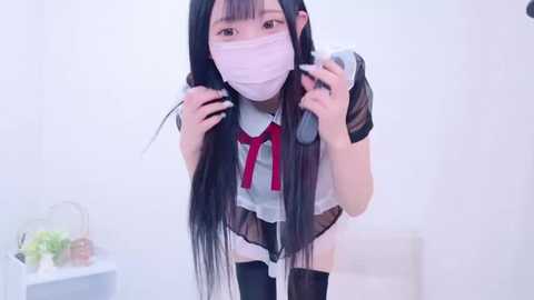 Media: Video of an East Asian woman with long black hair, wearing a white and red maid outfit, a face mask, and thigh-high stockings, posing with a camera in a bright, minimalist room.