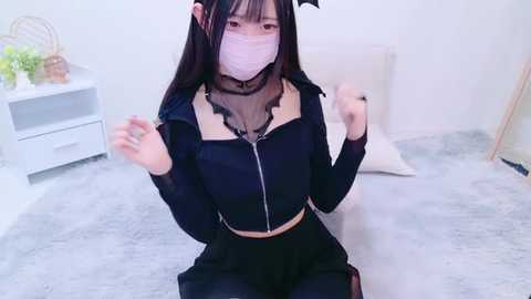 Media: Video of an Asian woman with long black hair, wearing a black and white bat-themed crop top, black pants, and a white mask, kneeling on a light carpeted floor.