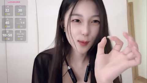 Media: Video of an East Asian woman with long black hair, wearing a black top and headphones, making an OK gesture with her right hand against a white wall.