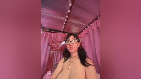 Media: A video of a topless East Asian woman with large breasts, black hair, and glasses, wearing a gold necklace, standing in a pink-tinted, draped bedroom.