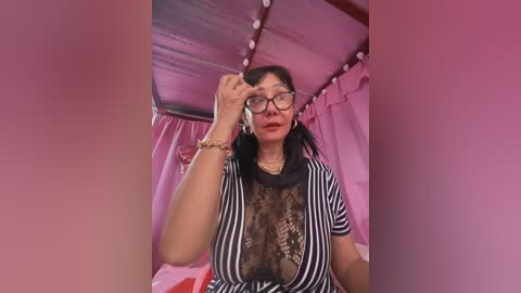 Media: A video of an Asian woman with glasses, black hair, and red lipstick, wearing a sheer black lace top over a striped shirt, adjusting her glasses in a pink-tinted room with string lights and heart-shaped decorations.