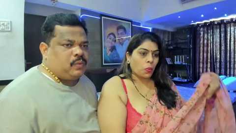 Media: Video of an Indian couple, a chubby man with a mustache and gold chain, and a woman with long dark hair and red saree, in a modern, well-lit living room with framed pictures and curtains.