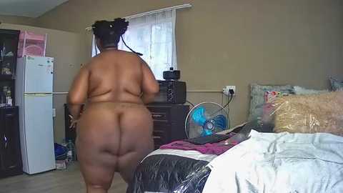 Media: Video of a large, dark-skinned woman with a round, curvy physique, wearing nothing, standing in a cluttered bedroom with a bed, white fridge, and fan.