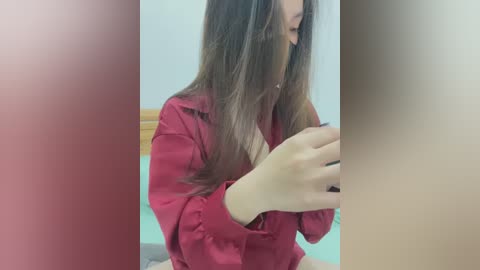 Media: Video of a young woman with long, straight brown hair, wearing a red satin robe, sitting on a bed, holding a smartphone, blurred background.