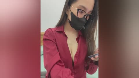 Media: Video of a young Asian woman with long dark hair, wearing a red button-down shirt exposing cleavage, black face mask, and glasses, holding a smartphone. Background is a blurred room with a wooden headboard.