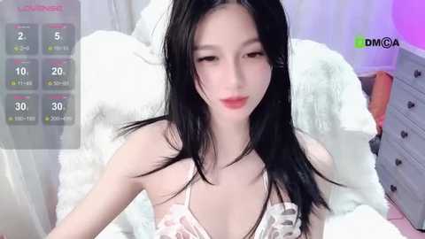 Media: A video of a young Asian woman with long black hair, pale skin, and red lips, wearing a white lace bra, sitting on a white fur blanket in a bedroom with purple walls and a dresser.