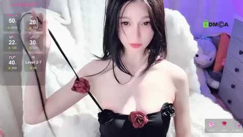 Media: A young Asian woman with fair skin and straight black hair poses in a bedroom, wearing a strapless black dress adorned with red roses. She holds a whip, standing against a white fur backdrop with plush toys and a green DMM.com watermark.