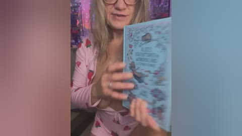 Media: Video of a blonde woman with glasses, wearing a pink floral robe, reading a comic book, in a colorful, sparkly room.