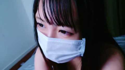 Media: A video of an Asian woman with long black hair and bangs, wearing a white medical mask, looking down. The background is dimly lit with a hint of a wall and floor.