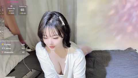 Media: Video of an East Asian woman with short black hair and fair skin, wearing a white top, lying on a bed in a dimly lit room, with a pink light effect in the background.