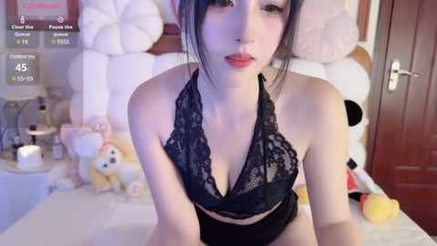 Media: Video of a young Asian woman with pale skin, dark hair, and full lips, wearing a black lace halter top, kneeling on a bed with plush toys and a wooden door in the background.