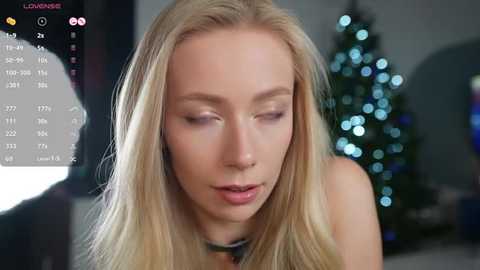 Media: A video of a blonde woman with fair skin and closed eyes, wearing a black choker, in a softly lit room with a blurred Christmas tree in the background.