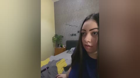 Media: Video of a young woman with long dark hair, wearing a blue shirt, peeking through a doorway into a messy bedroom with a gray bed, yellow blanket, and green potted plant.