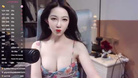 Media: Video of an East Asian woman with fair skin and long black hair, wearing a low-cut floral dress, seated in a dimly lit room, using a webcam.