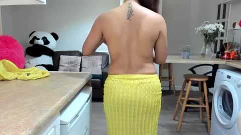 Media: Video of a topless woman with a tattoo on her back, wearing a yellow knitted skirt, standing in a modern, minimalist kitchen with a panda plush, white flowers, and a washing machine.