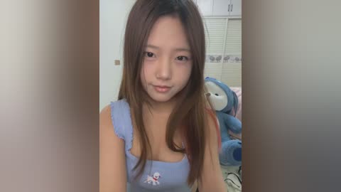 Media: Video of an Asian woman with straight, long brown hair, wearing a blue sleeveless top with a cartoon character, standing in a bedroom with a stuffed bear and white blinds in the background.