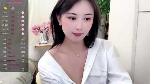 Media: Video of an East Asian woman with fair skin, straight black hair, and a slender physique. She wears a white blouse, a dainty necklace, and sits indoors with a blurred background of flowers and a green plant.