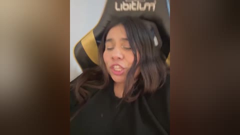 Video of a young woman with medium brown skin and long, dark hair, wearing a black top, sitting in a black and yellow gaming chair, with her eyes closed and mouth slightly open, suggesting a relaxed or sleepy state.