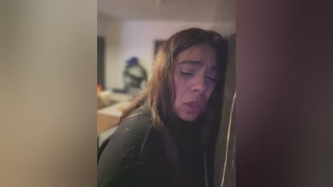 Video of a young woman with long brown hair, wearing a black sweater, leaning against a wooden door, eyes closed, expression sad, blurred background.