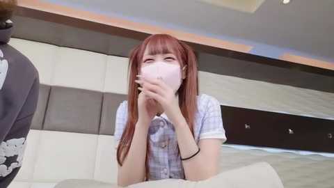 Media: Video of a young Asian woman with straight, shoulder-length red hair, wearing a white face mask, plaid shirt, and black bracelet, seated indoors on a beige couch, with a person in a dark floral shirt partially visible.