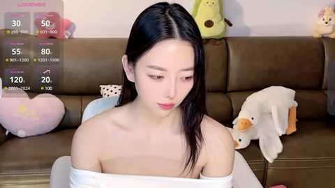 Media: A video of an East Asian woman with straight black hair, wearing a white off-shoulder top, sitting on a brown leather couch. Background features plush toys, including a yellow duck and a heart-shaped pillow.